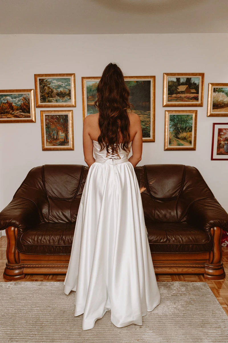 Mladenka u bijeloj satenskoj vjenčanici stoji pred smeđom kožnom sofom, promatrajući galeriju pejzažnih slika..