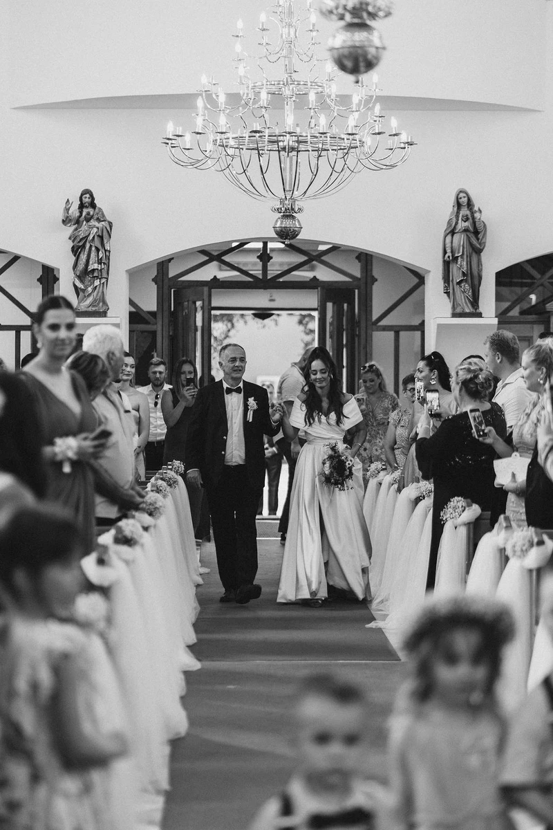 Mladenka hoda prema oltaru, uz starijeg gospodina, dok gosti gledaju i fotografiraju u prekrasno ukrašenom prostoru...