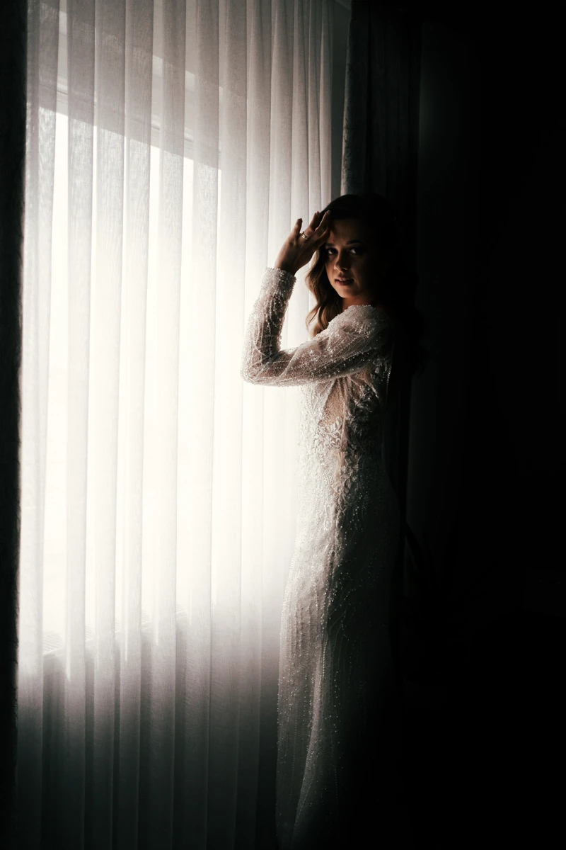 Mladenka u vjenčanici stoji uz prozor, obasjana toplim dnevnim svjetlom, spremna za izlazak iz kuće na svoj svadbeni dan.