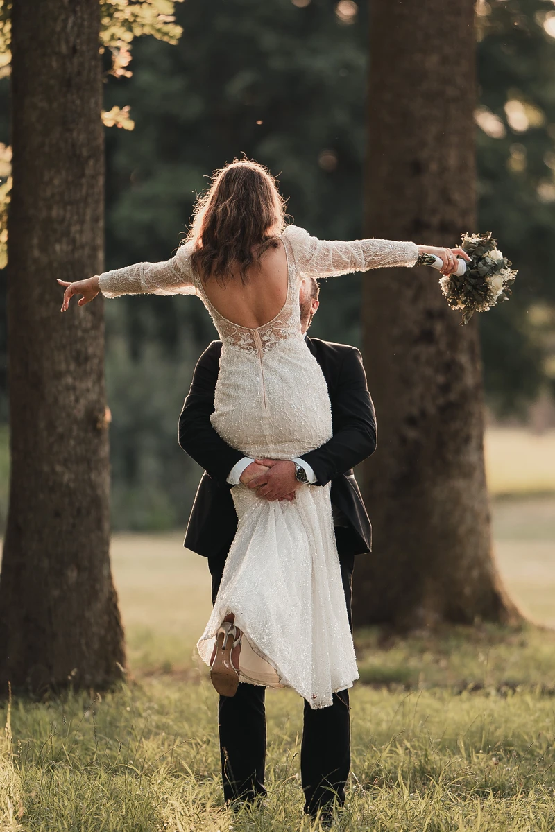 Mladoženja na travi podiže mladenku s raširenim rukama i buketom, okruženi slavonskom prirodom i šumama.