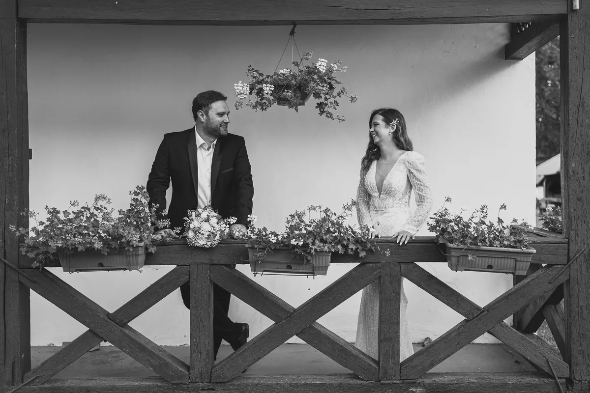 Crno-bijela fotografija mladenaca koji stoje kraj stare kuće i drvene ograde u slavonskom sokaku.